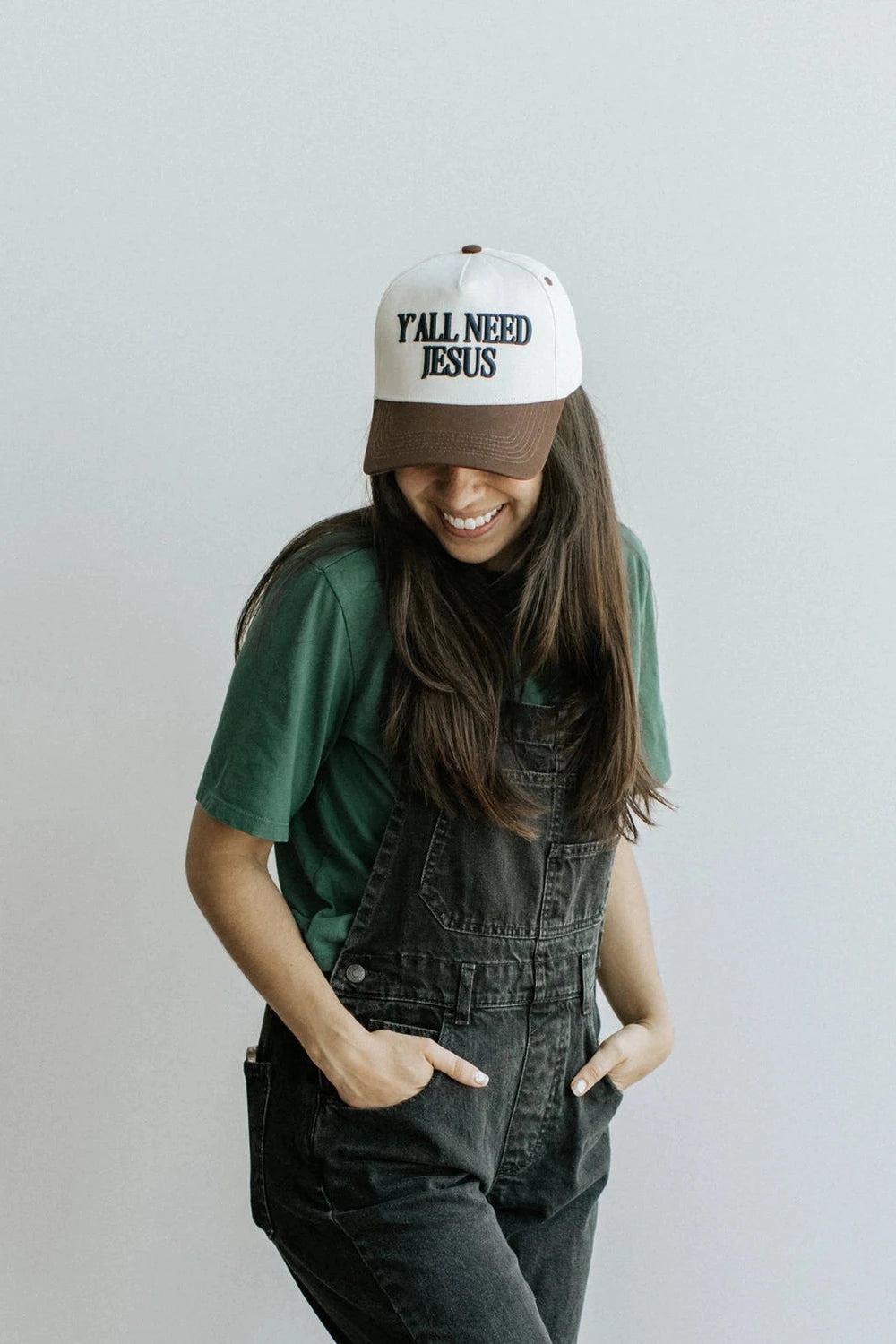 Person wearing a cap with 'Yall Need Jesus' text, green shirt, and black overalls on a plain background