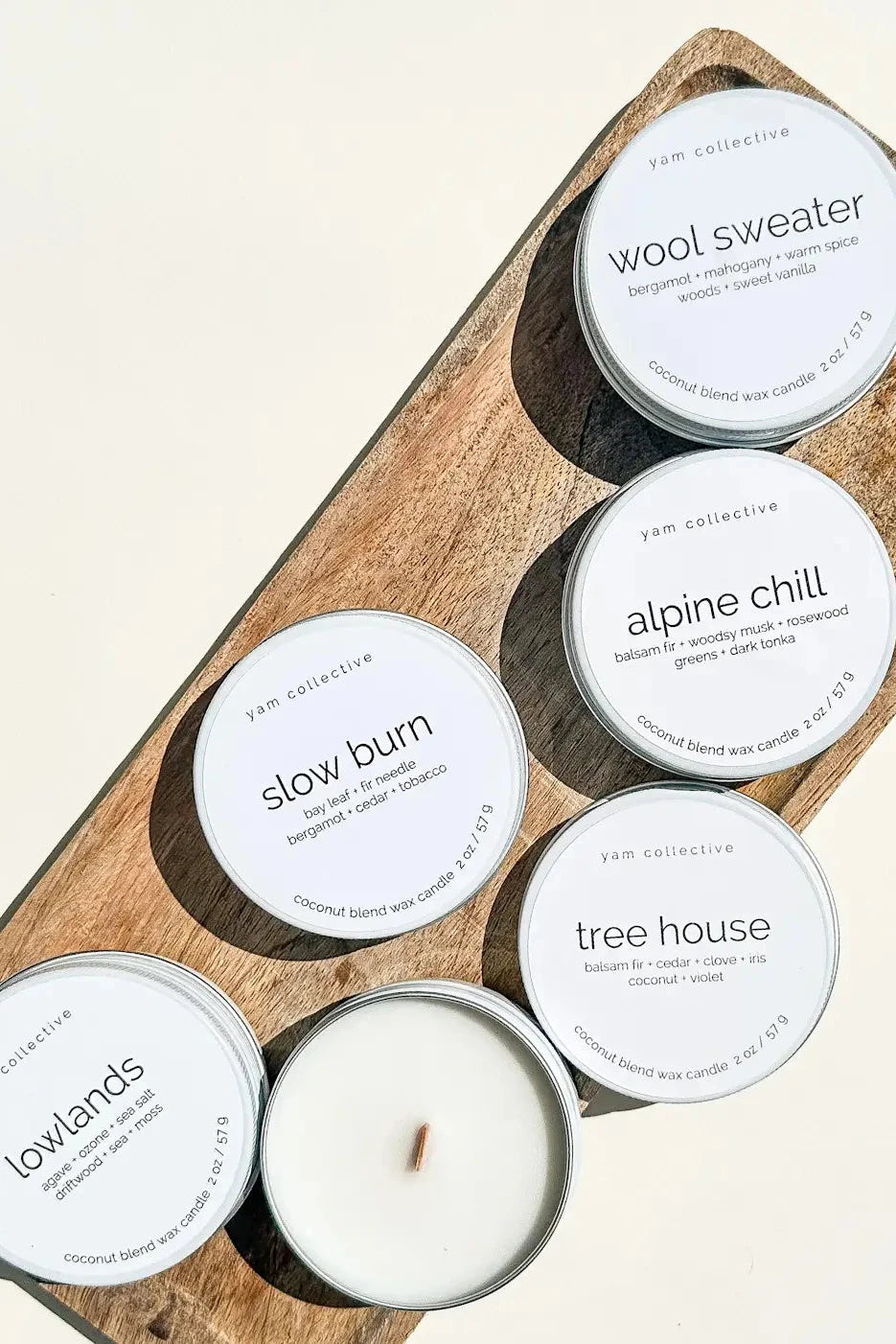 Five scented candles from the brand Pap Collection are arranged on a rectangular wooden tray against a warm off-white background. One candle is open, showing white wax and a single wick, while the others are closed with white labels.