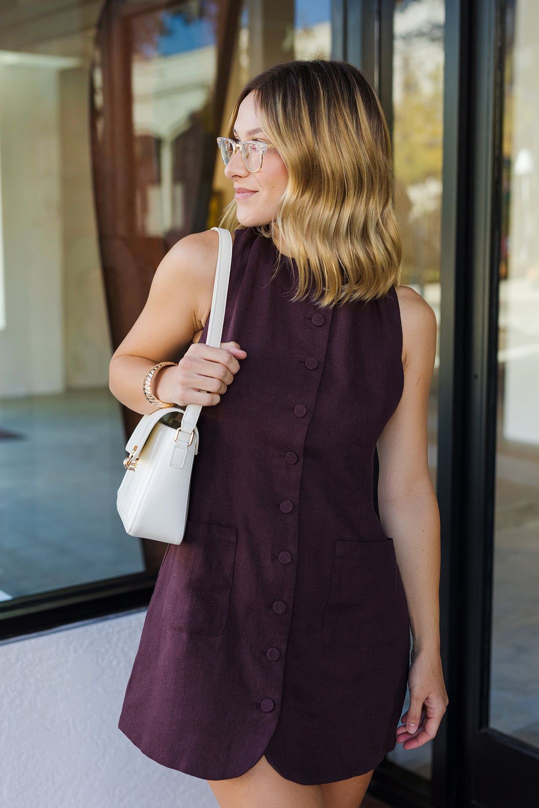 Woman in a dark sleeveless dress holding a white handbag, standing in front of glass doors.